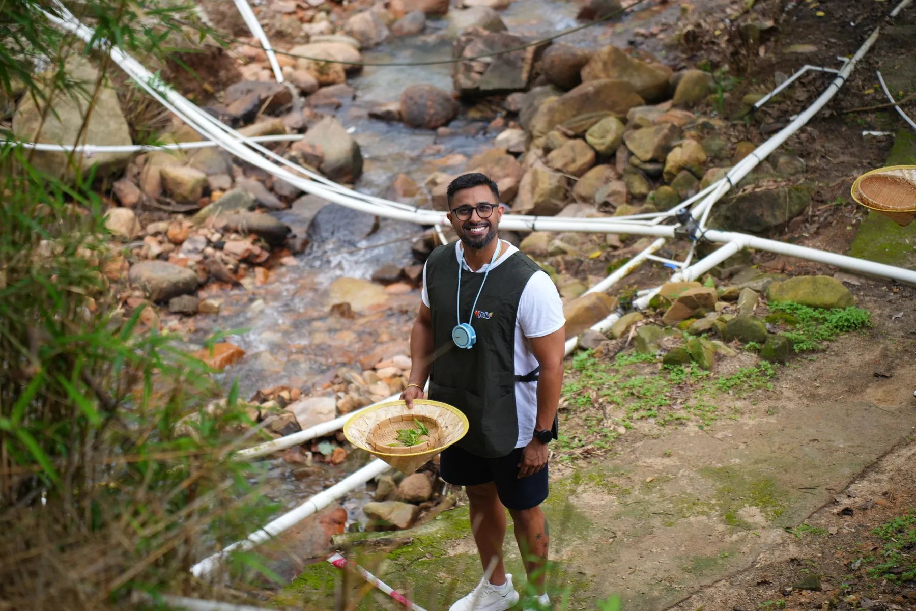 Dhee grinning beside a mountain stream, holding a bamboo basket of fresh tea leaves.